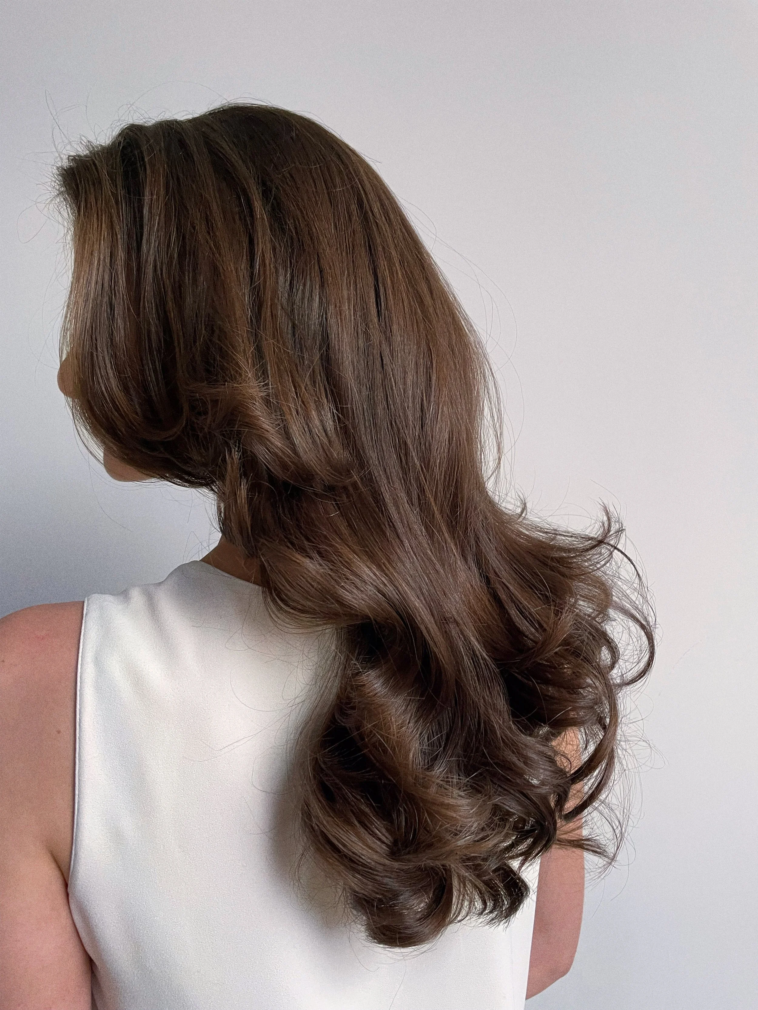 Woman with long, wavy brown hair wearing a white top, photographed from the side against a light grey background.