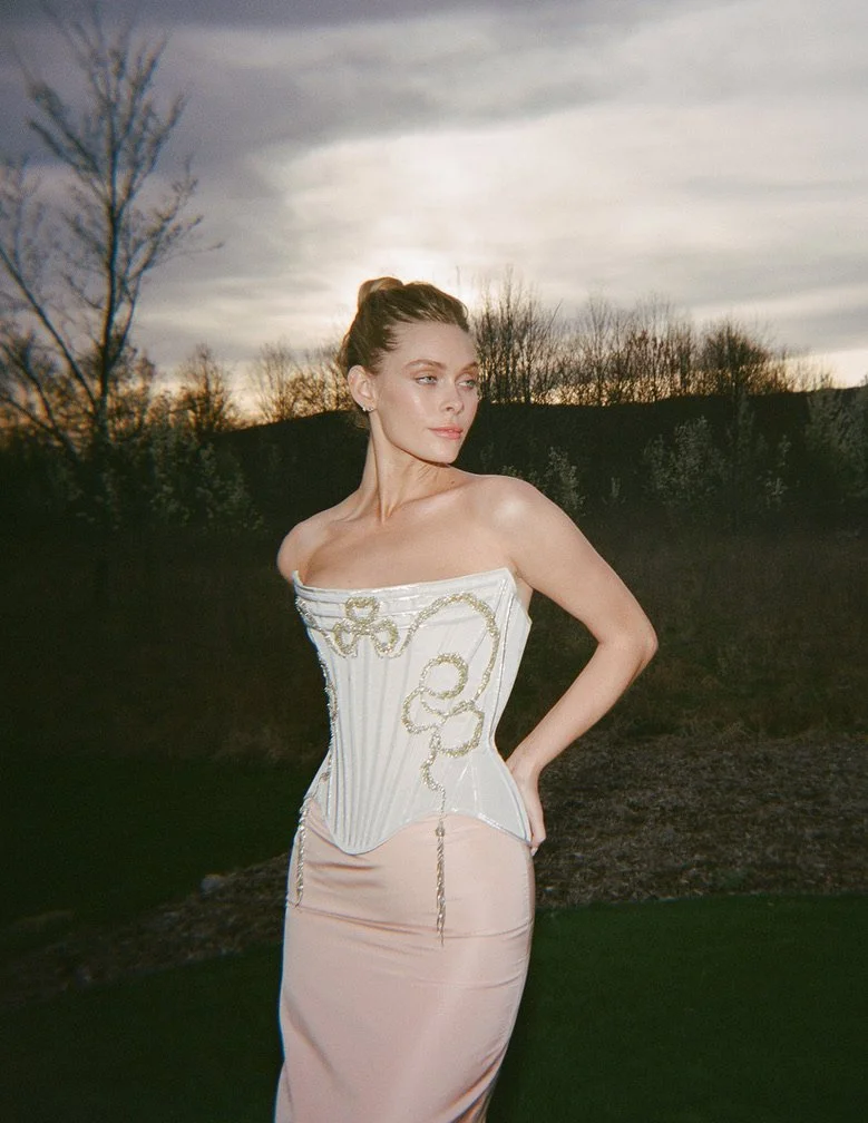 A woman standing outdoors at sunset, wearing an elegant strapless dress with a white and gold corset top and a light pink satin skirt. She has an elaborate updo and appears to be gazing thoughtfully into the distance.