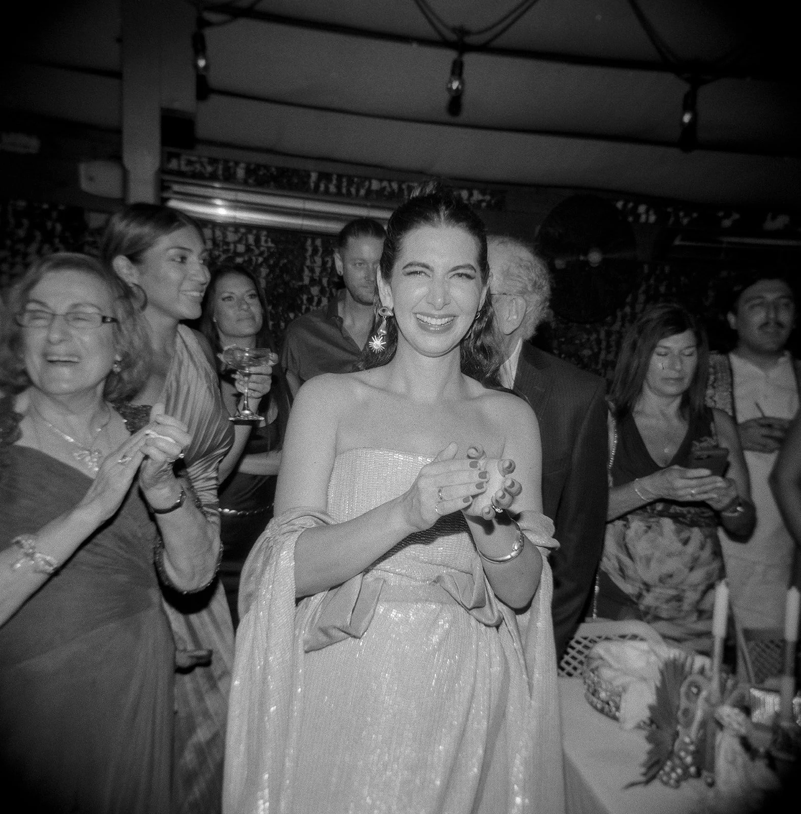 A group of people, mostly women, at a party, with one woman smiling and clapping in the center, wearing a strapless dress and earrings.