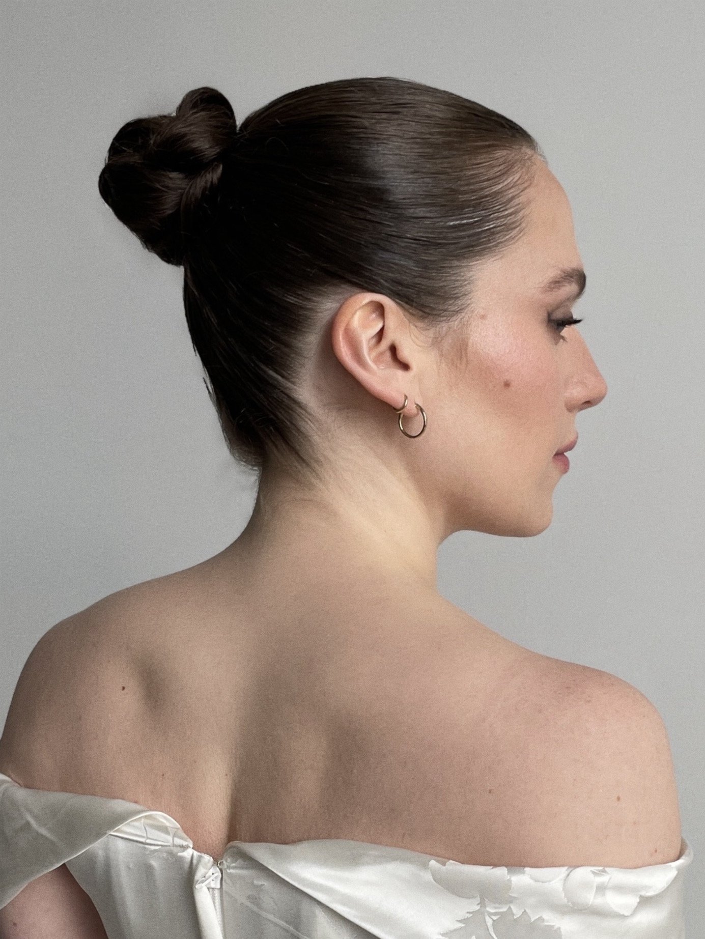 Portrait of a woman with dark brown hair styled in a sleek bun, wearing small earrings, facing to the right, with her shoulder and part of her upper back exposed, against a plain light gray background.