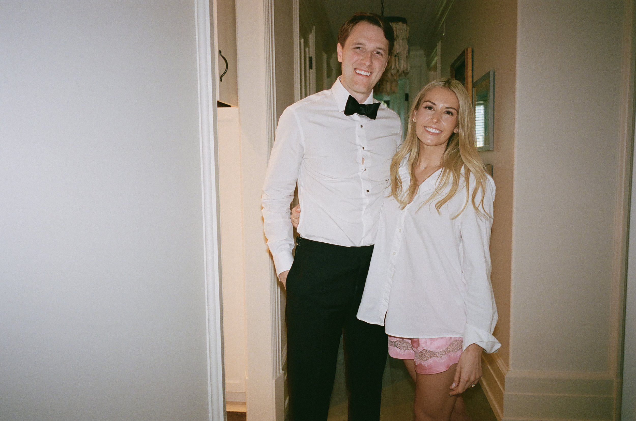 A man and woman standing together indoors, smiling at the camera. The man is dressed in a white shirt with a black bow tie and black pants, and the woman is wearing a white shirt and pink shorts.