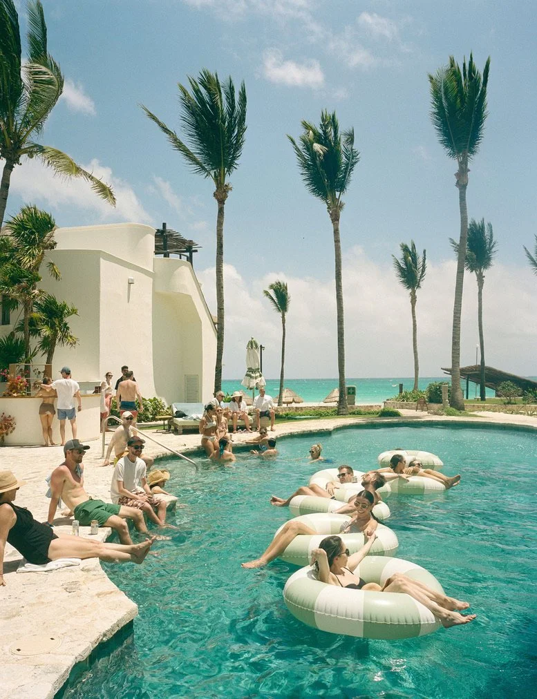 People relaxing in a swimming pool surrounded by palm trees at a beach resort on a sunny day.