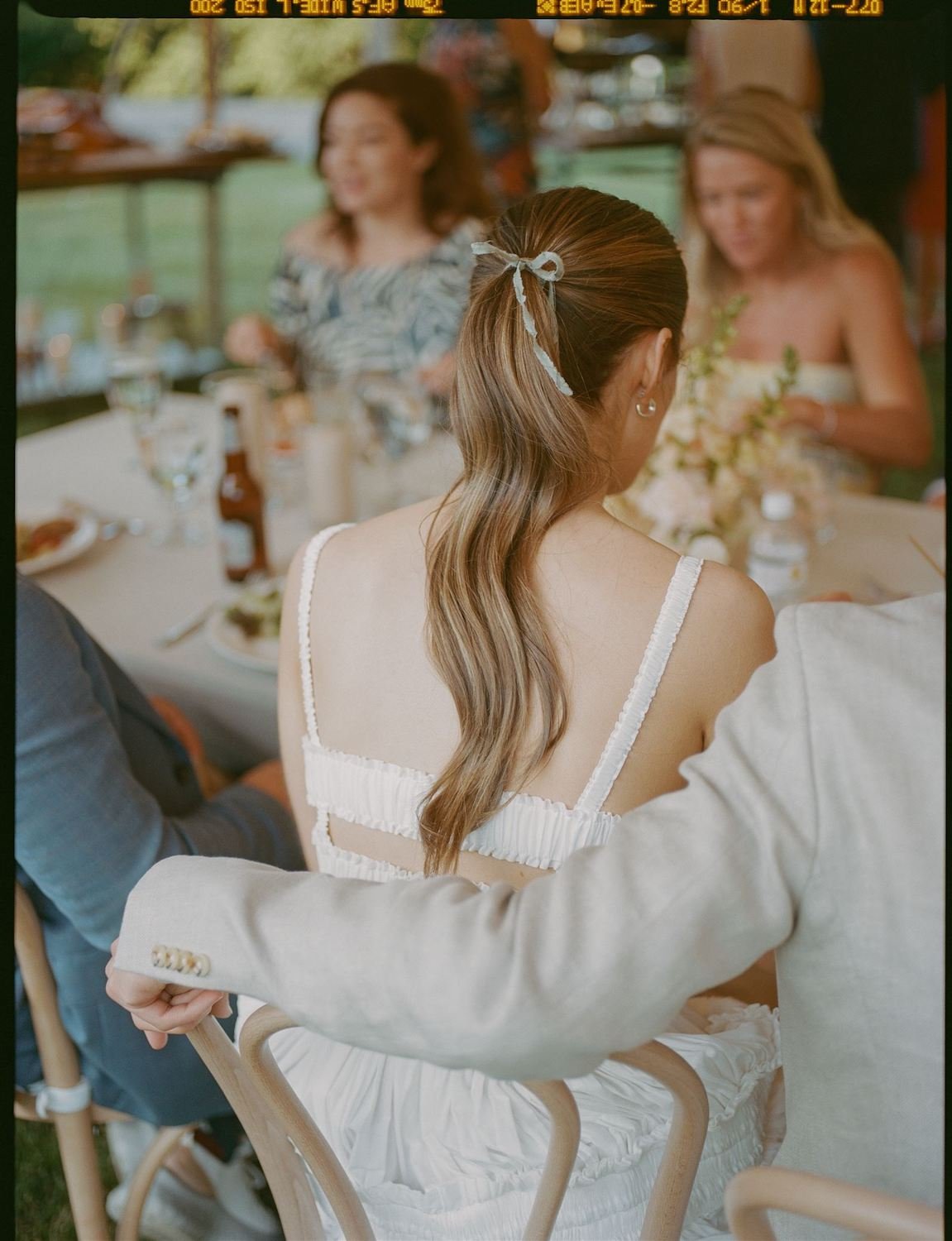 A woman with long, wavy, light brown hair tied with a white ribbon sits at a table during a social gathering. She is seen from the back and wears a white dress with thin straps. There are other women and people around the table, with glasses, plates, and bottles visible, indicating a meal or celebration.