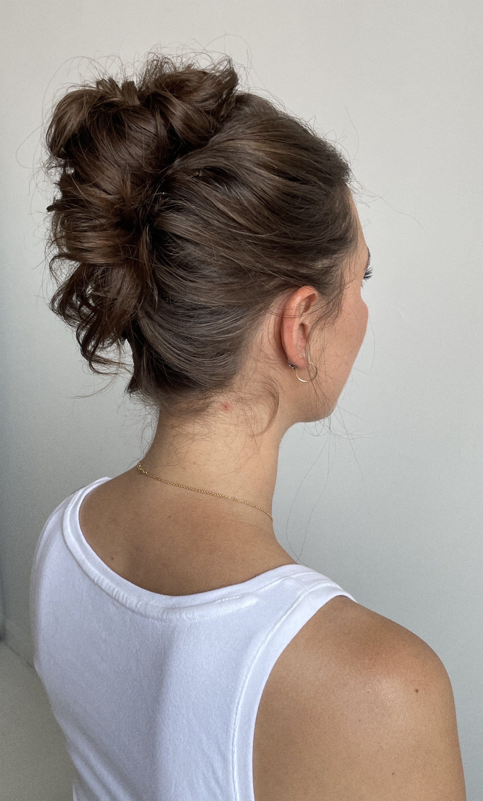Back view of a woman with brown hair styled in a messy bun, wearing a white sleeveless top, gold hoop earrings, and a delicate gold necklace.