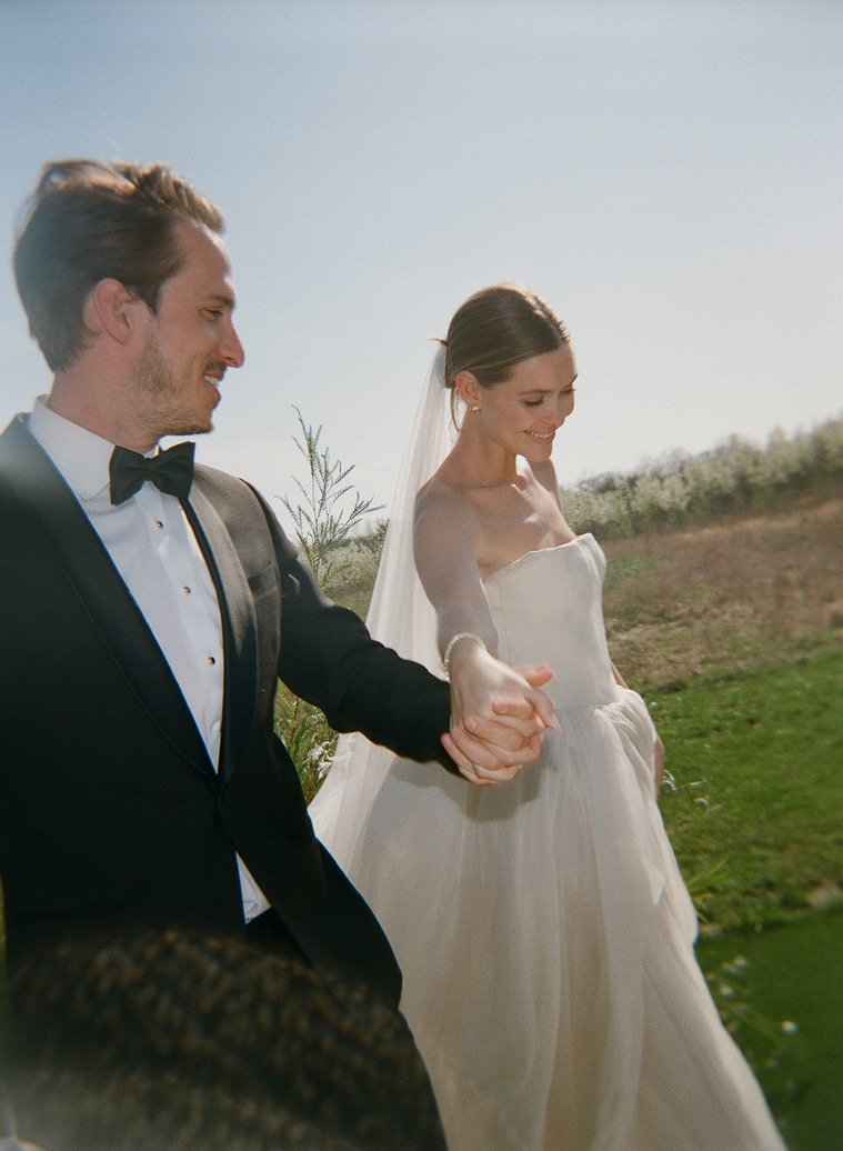 A bride and groom holding hands outdoors on a sunny day, both smiling.