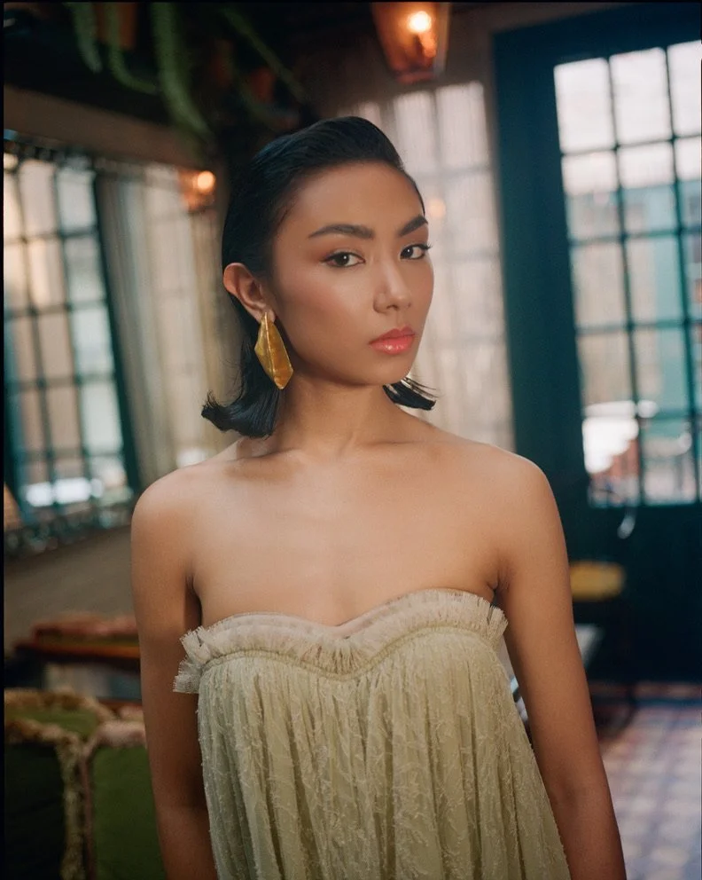 A woman with dark, shoulder-length hair, wearing large yellow earrings and a strapless beige dress, stands inside a room with large windows and colorful decorations.