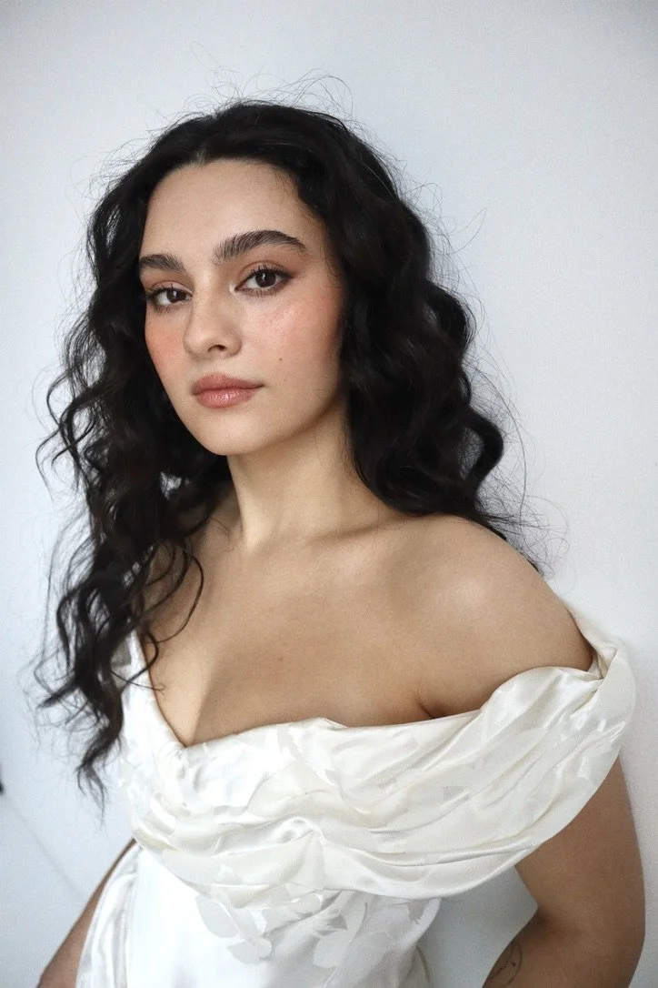 A young woman with long, curly black hair and light skin wearing a white, off-the-shoulder dress.