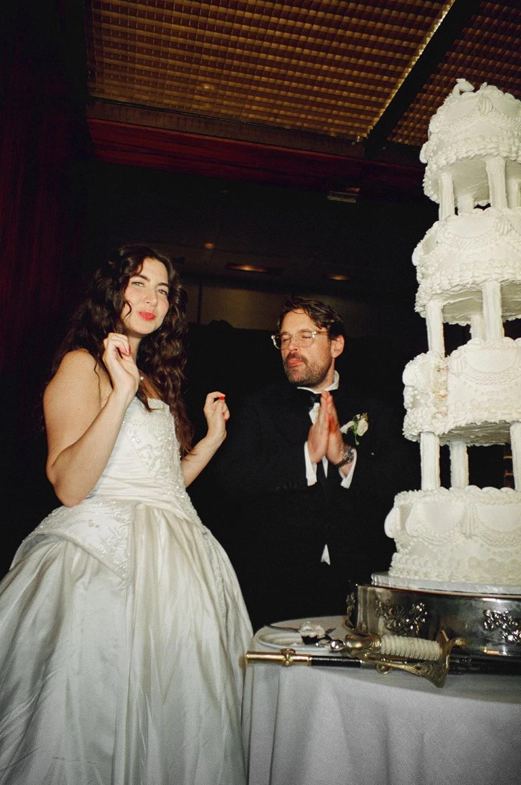 A bride in a white wedding dress and a groom in a tuxedo pose near a large three-tiered white wedding cake at a wedding celebration.