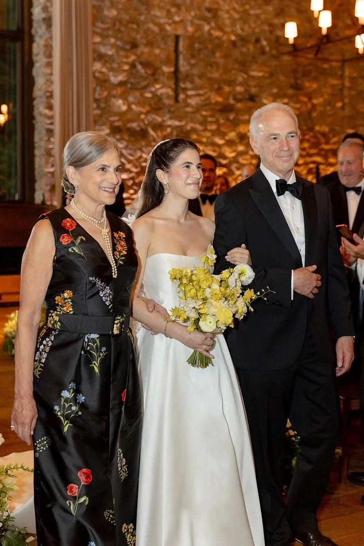 A young woman in a white wedding dress holding a bouquet of yellow and white flowers, walking down the aisle with her parents at a wedding ceremony in a warmly lit venue with a stone wall background.