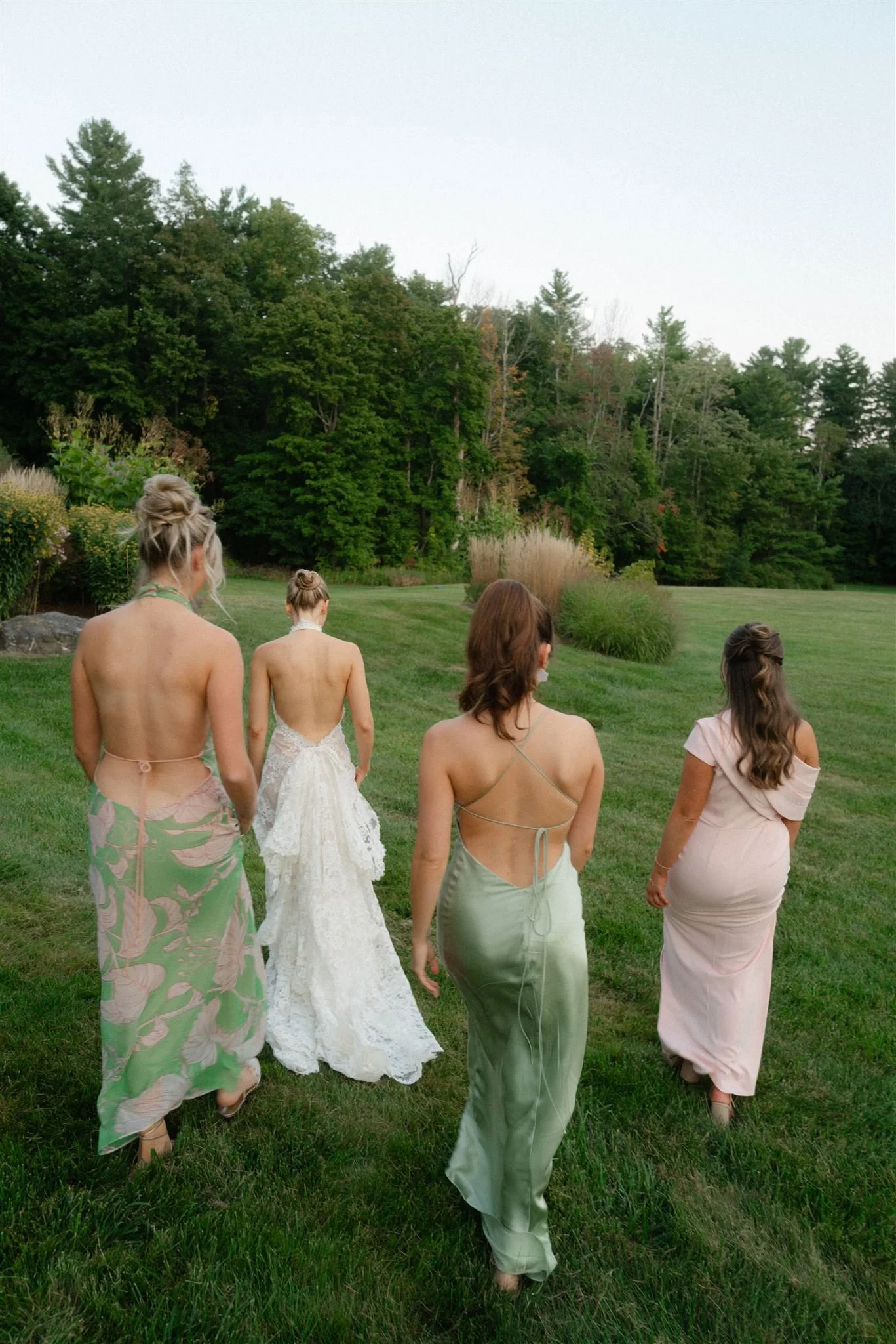 Four women in elegant dresses walking on a grassy field with trees in the background.
