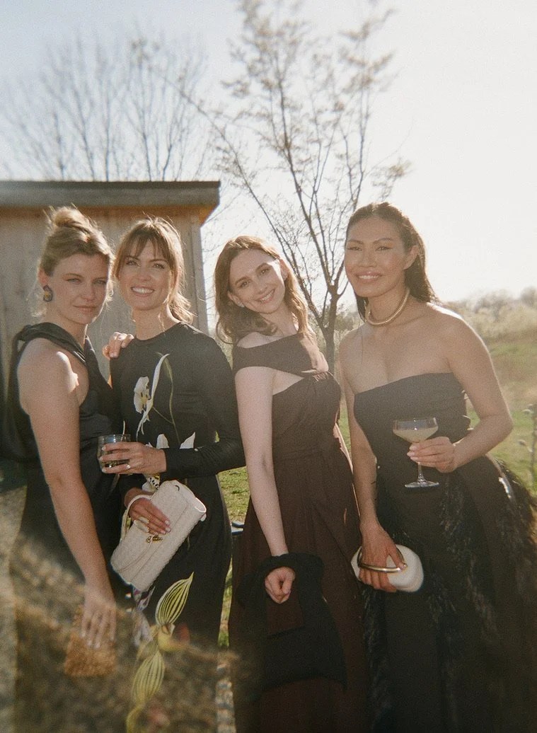 Four women in elegant dresses standing outdoors, smiling and holding drinks during daytime.
