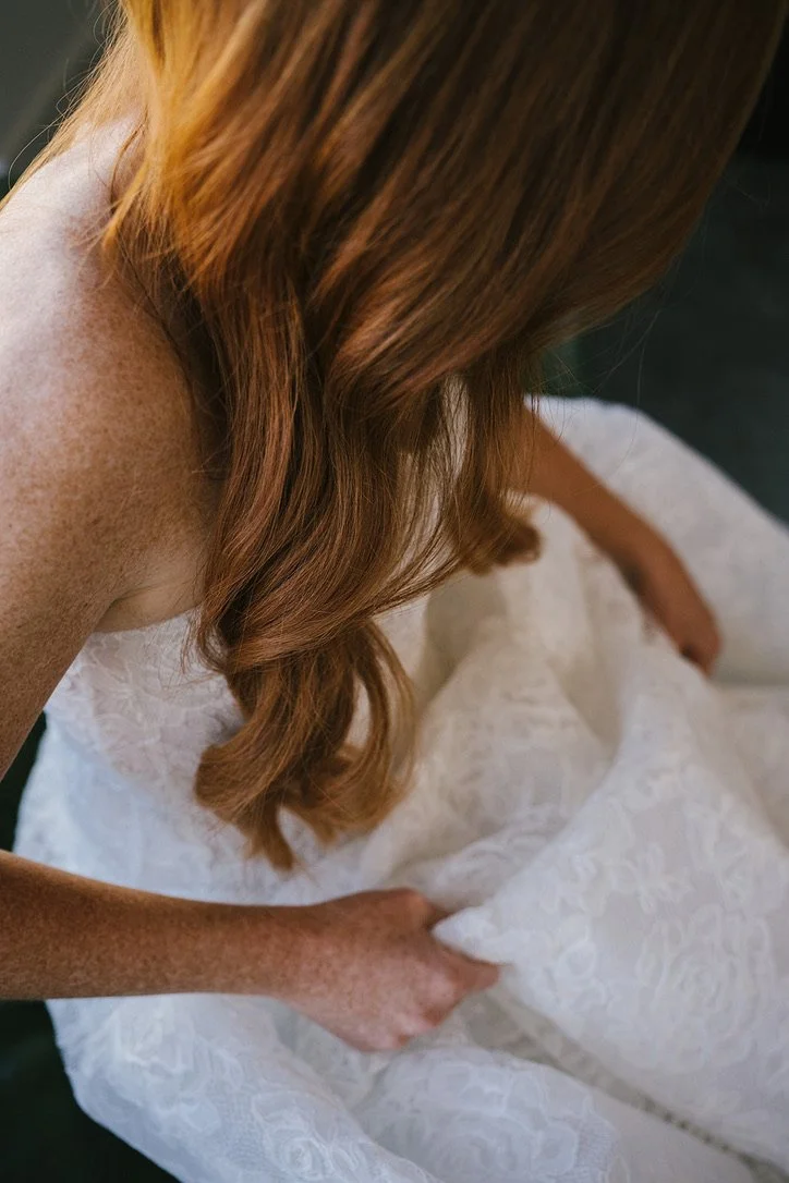 A woman with long, wavy red hair adjusts her white lace dress.