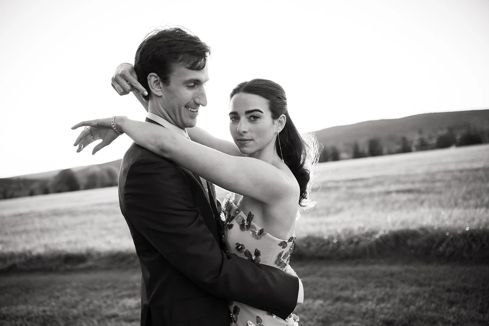 Black and white photo of a couple embracing outdoors, the man in a suit and the woman in a dress with butterfly details, with a countryside landscape in the background.