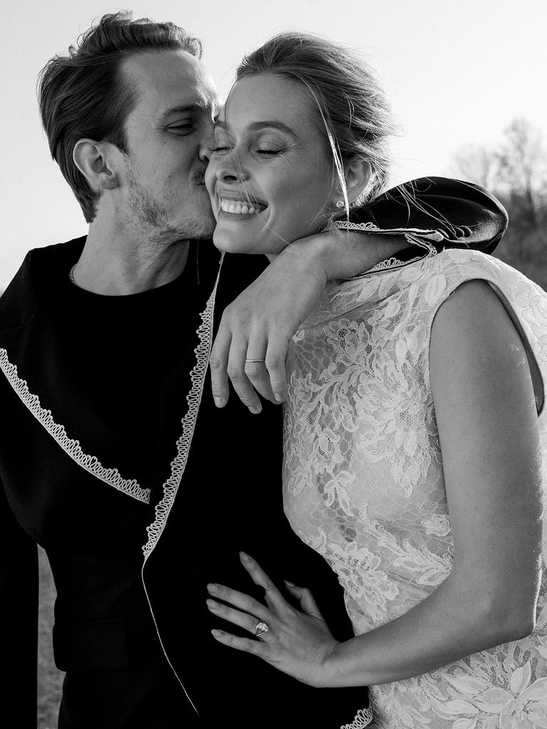 A couple on their wedding day sharing a tender moment, with the man kissing the woman's cheek and the woman smiling with her eyes closed, outdoors in natural light.
