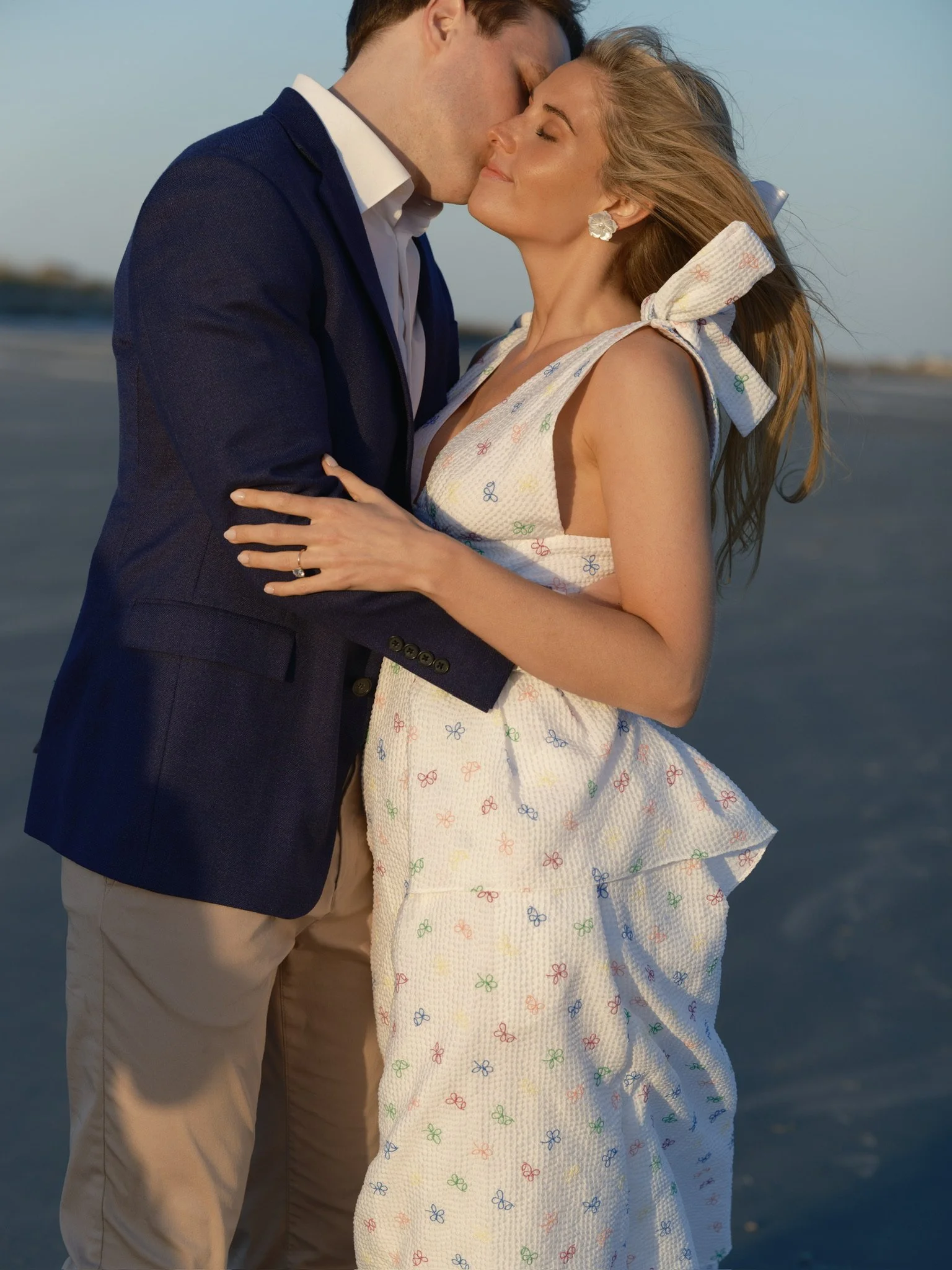 A couple is kissing on the beach during sunset. The man is wearing a navy blazer and khaki pants, and the woman is wearing a white dress with colorful small bow prints and a large bow on her shoulder.