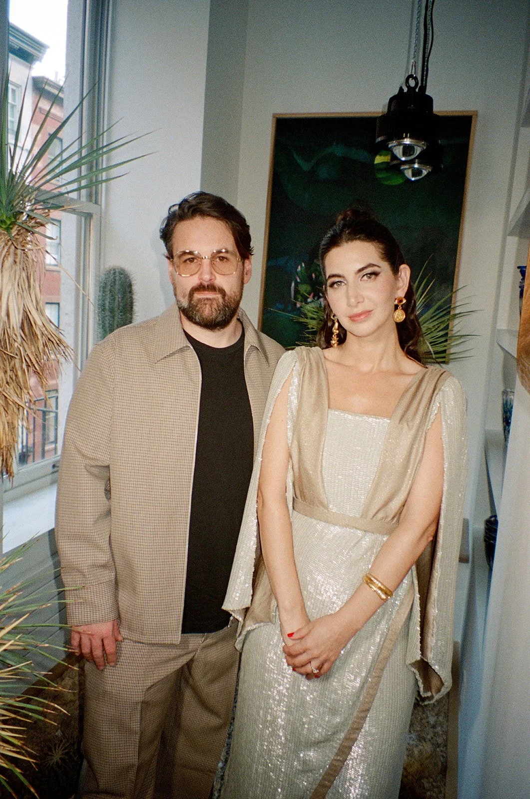 A man with glasses and a beard wearing a beige jacket and black shirt, standing next to a woman in a shiny, cream-colored gown with gold accents, accessorized with gold earrings and bracelets, in an indoor space with plants and artwork in the backgro
