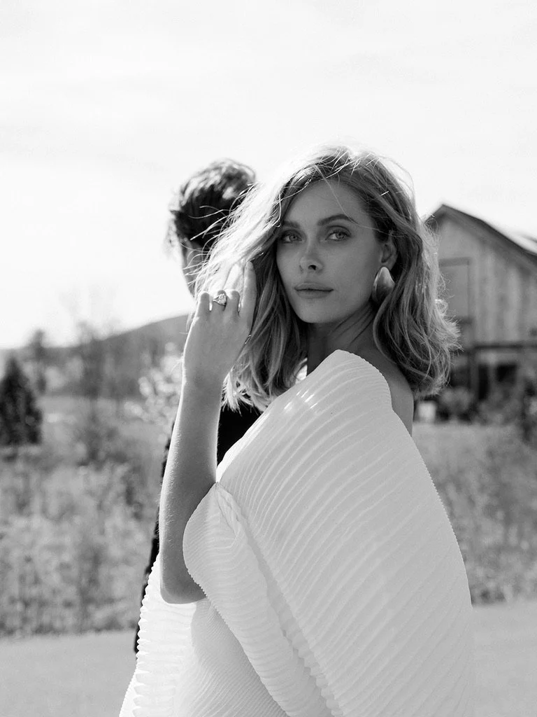 A black and white photo of a woman with shoulder-length hair, wearing a textured white top, standing outdoors with a blurry background of a wooden house and trees.