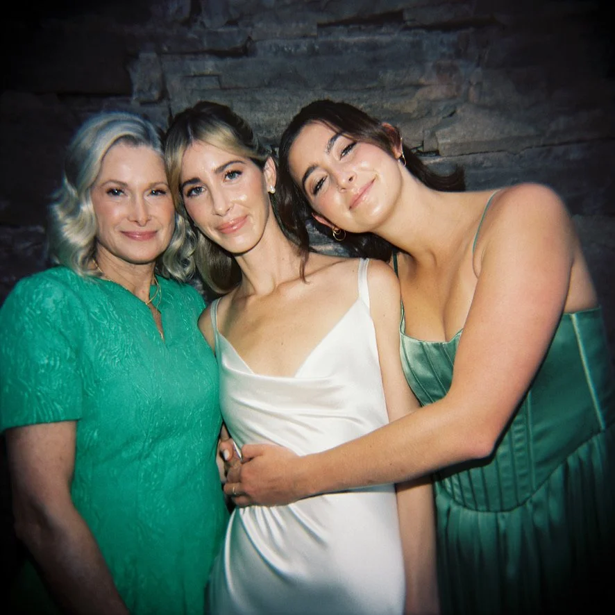 Three women standing close together, smiling, dressed in elegant attire, against a stone wall background.