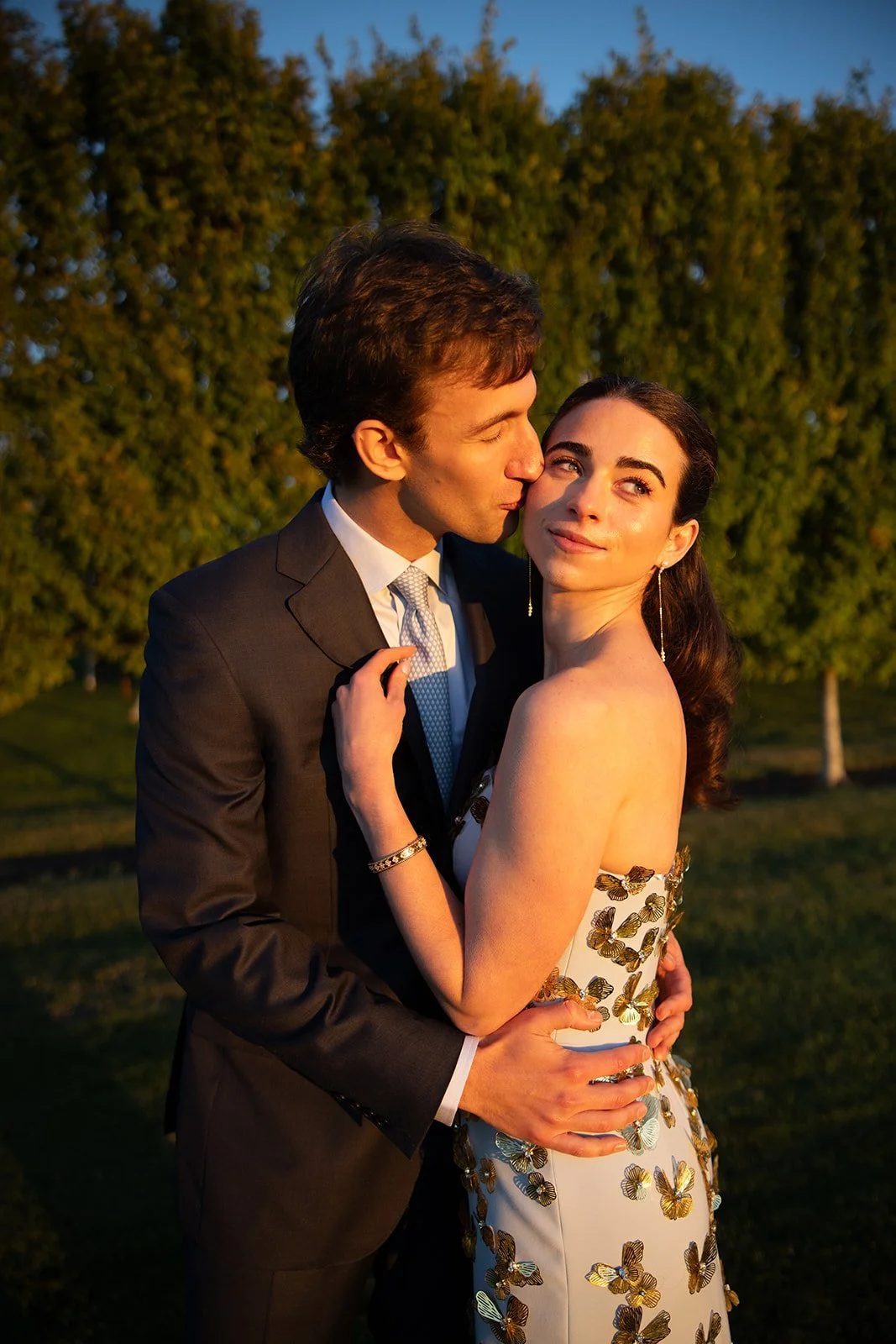 A romantic scene with a man in a black suit kissing a woman in an elegant dress with butterfly embellishments on a grassy field at sunset.