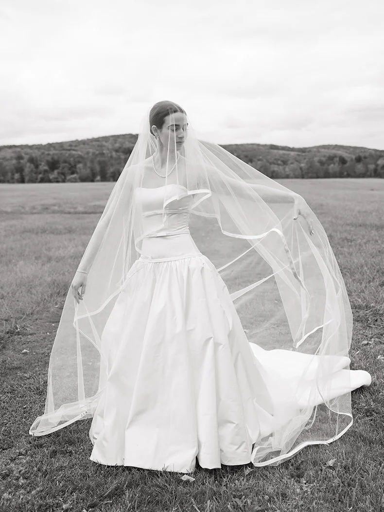 A woman in a wedding dress standing outdoors in a field with rolling hills in the background, holding part of her veil.