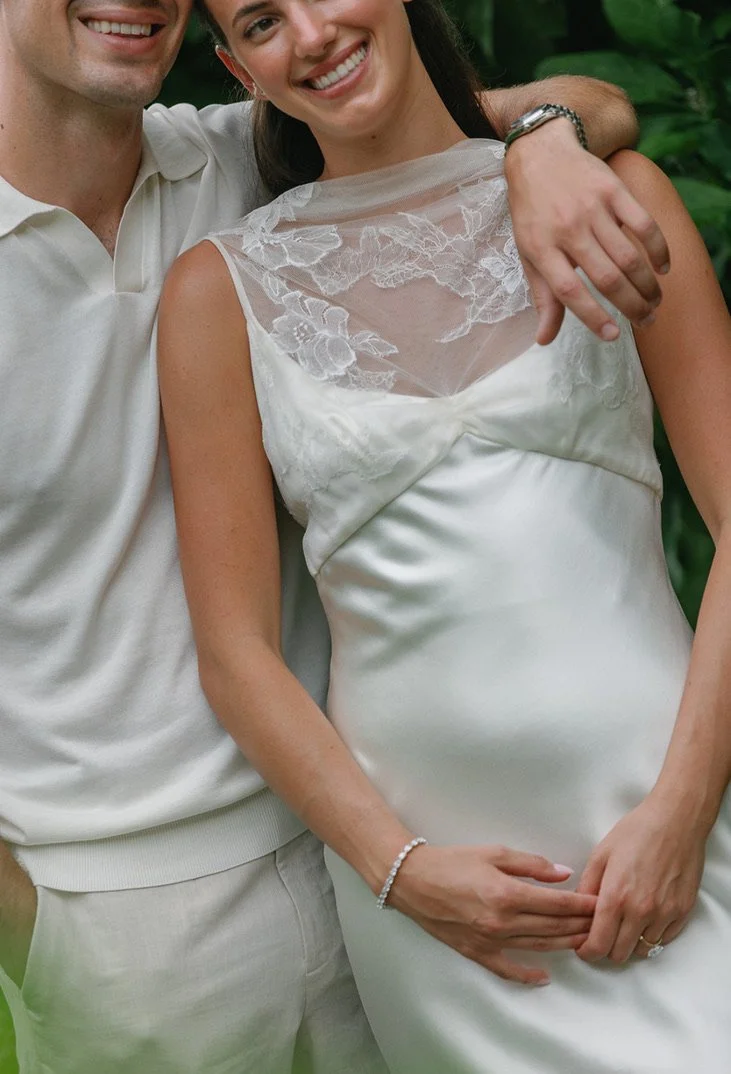 A smiling woman in a white dress with lace details and a man in a white shirt, standing close together outdoors with green foliage in the background.