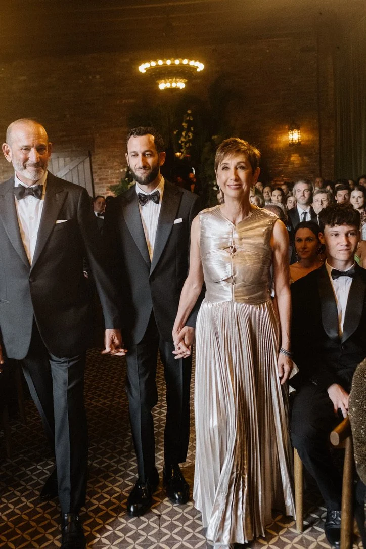 Group of elegantly dressed people at a formal event, holding hands and standing in a row with an audience in the background.