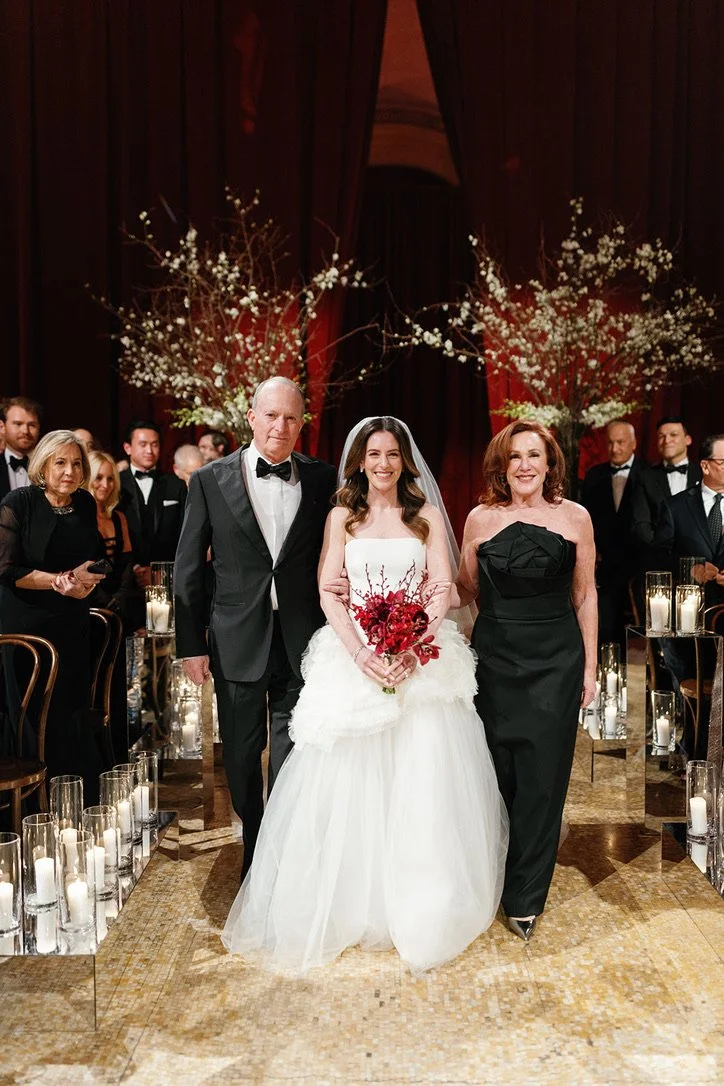 Bride walking down the aisle with her parents during a wedding ceremony in an elegant indoor venue decorated with candlelit lanterns and floral arrangements.