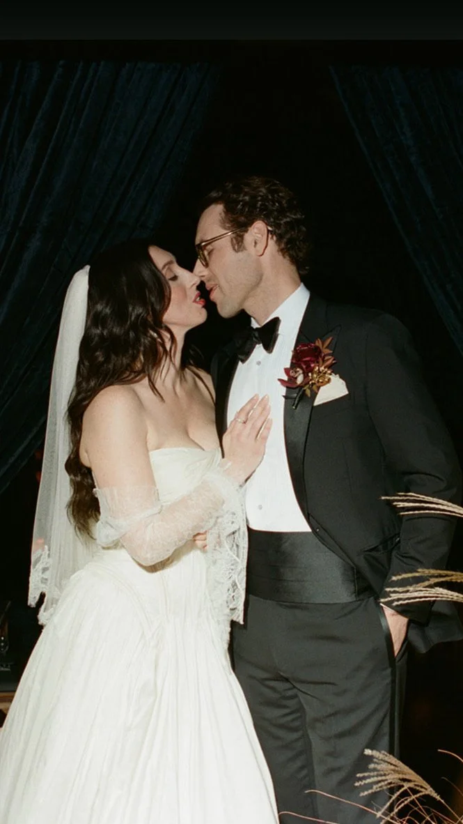 A bride and groom sharing a close moment at their wedding, with the bride wearing a white strapless gown and veil, and the groom in a black tuxedo with a boutonniere, in front of dark curtains.