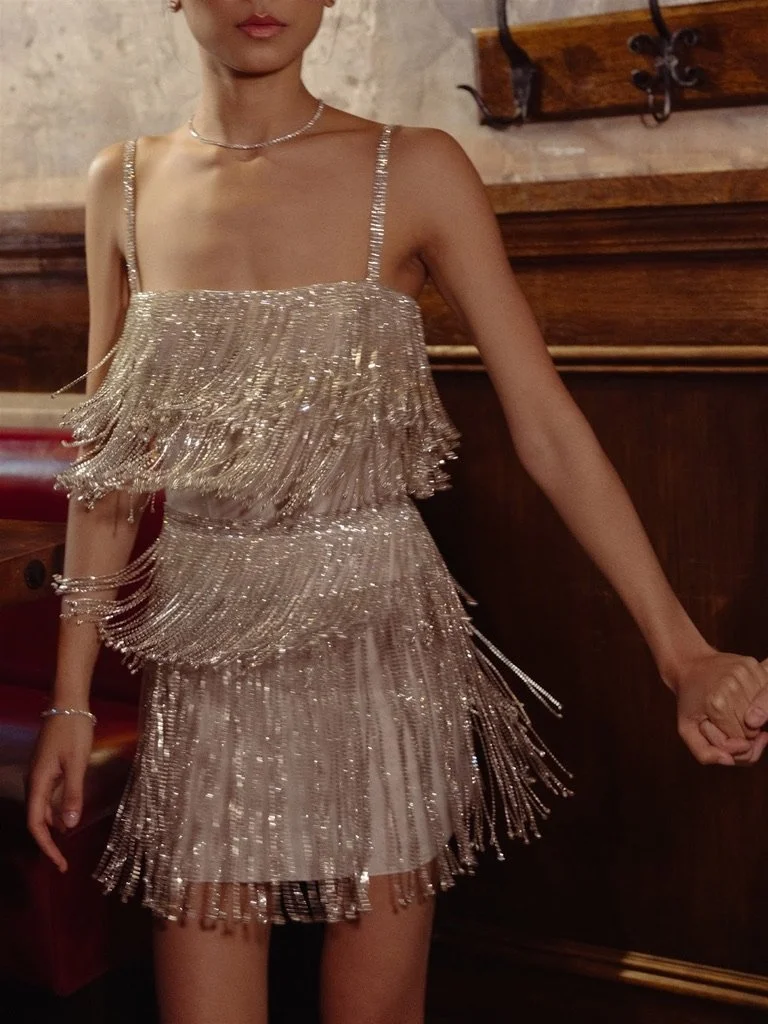 Woman wearing a sparkly, layered, fringe dress with thin straps and jewelry, in a cozy indoor setting.