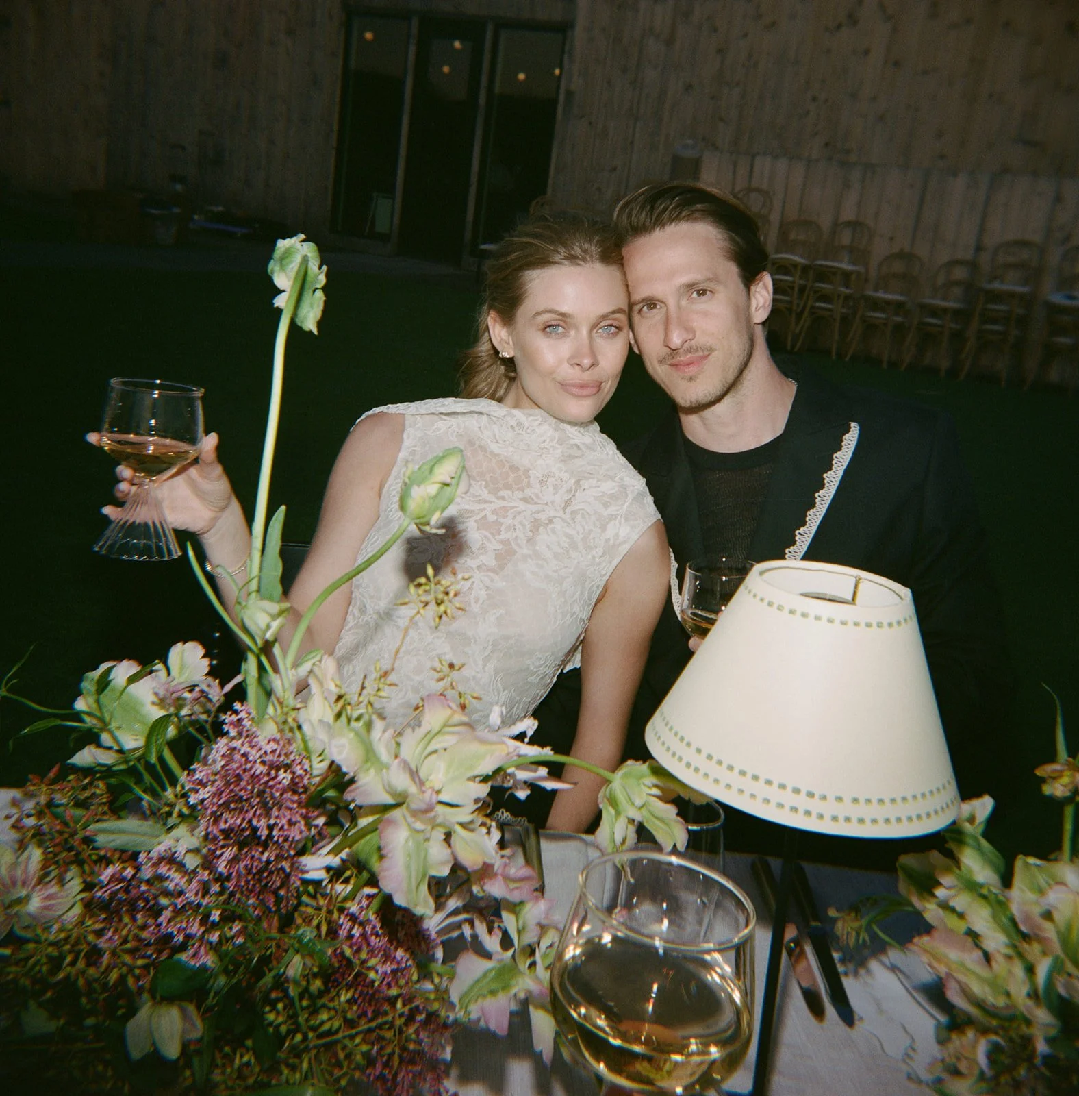 A woman and a man dressed up at a dinner event, sitting closely together. The woman is holding a glass of wine, and several glasses are visible. There is a floral centerpiece and a table lamp with a white shade in front of them.