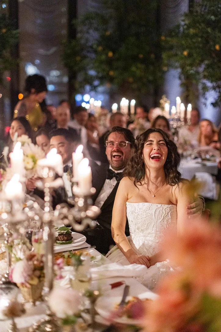People laughing and enjoying a wedding reception at a decorated banquet table with candles and flowers, outdoors at night.