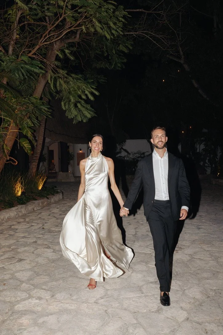 A romantic couple walking hand-in-hand outdoors at night, surrounded by trees and soft lighting, dressed in formal attire — the woman in a flowing, satin evening gown and the man in a dark suit.