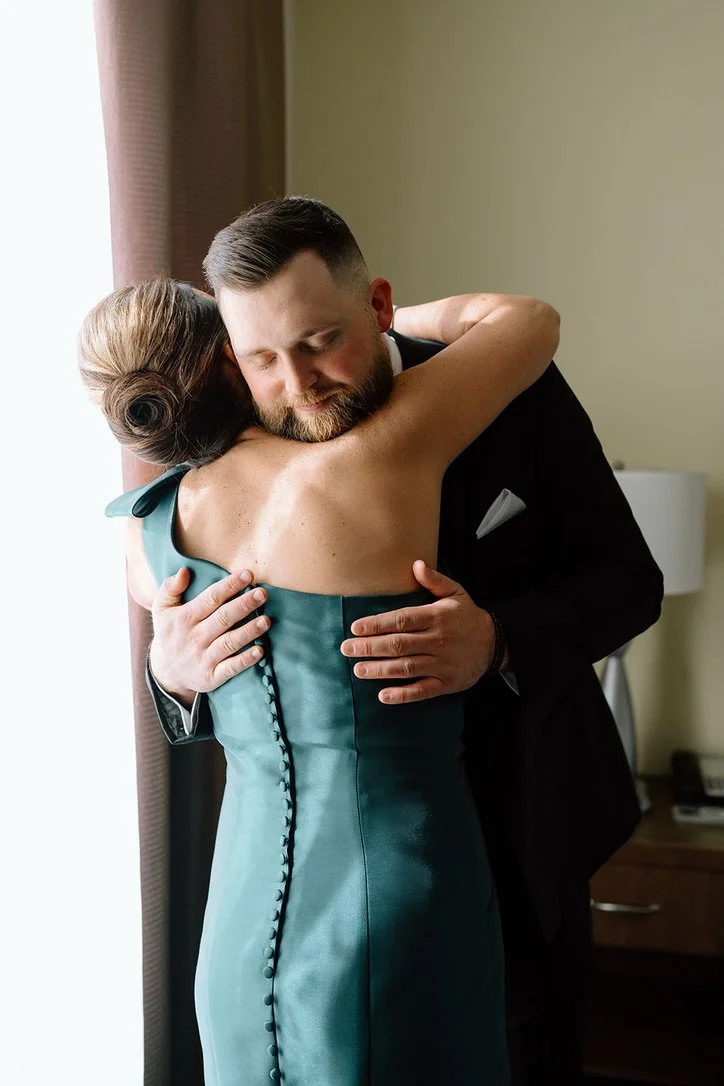 A couple hugging, the woman wearing a teal dress and the man in a dark suit, standing near a window in a room.