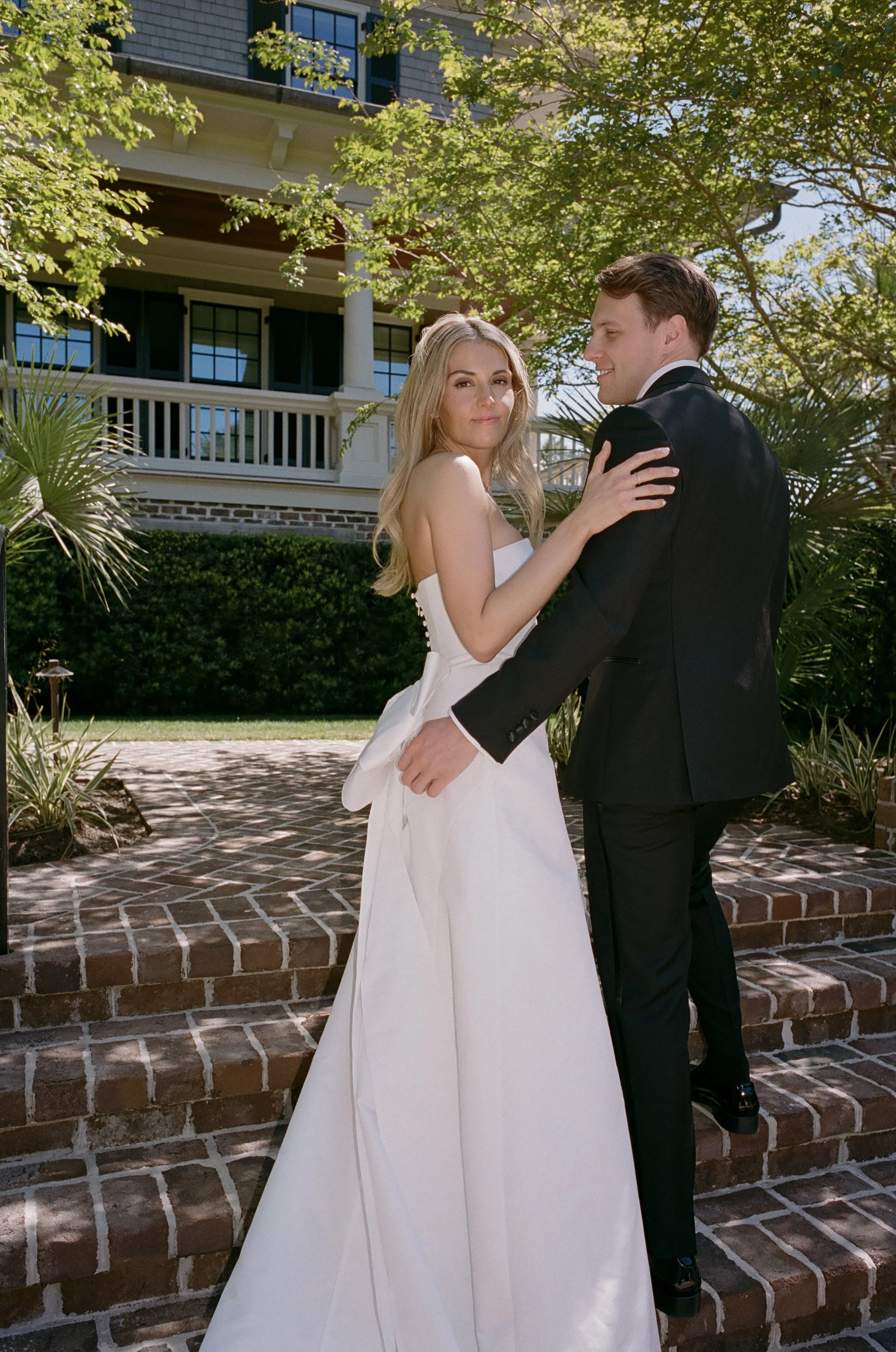 A couple dressed in wedding attire dancing outdoors on brick steps, with lush greenery and a house in the background.