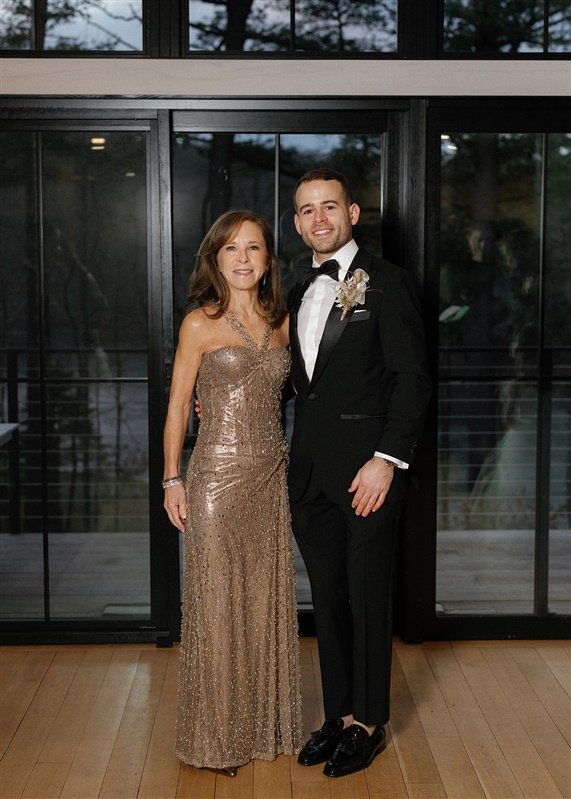A woman in a gold, sparkly evening gown standing next to a man in a black tuxedo with a bow tie, both smiling inside a modern building with large glass windows and a wooden floor.