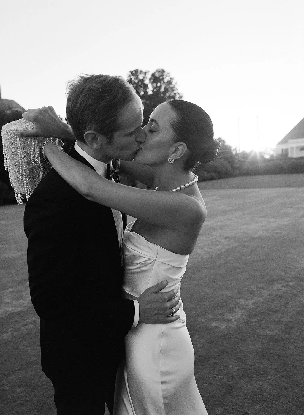 A black and white photograph of a bride and groom kissing outdoors during sunset. The bride wears a strapless wedding dress, pearl necklace, and earrings. The groom wears a tuxedo with a bow tie. The bride holds a clutch purse with pearl embellishmen