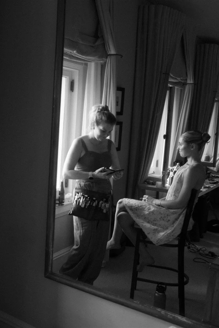 A woman sitting on a high chair in a room, looking straight ahead, while another woman stands in front of her near a window, looking at her phone, with makeup tools on a table nearby.