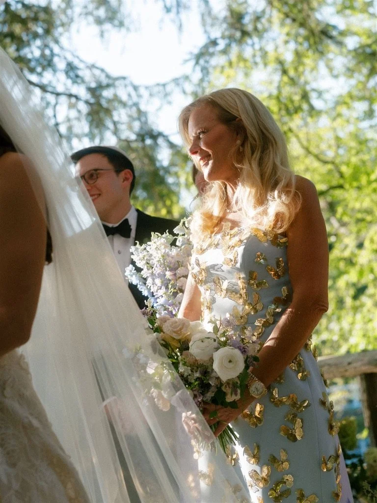A woman in a white dress with gold butterfly embellishments holding a bouquet of white and purple flowers at an outdoor wedding ceremony.