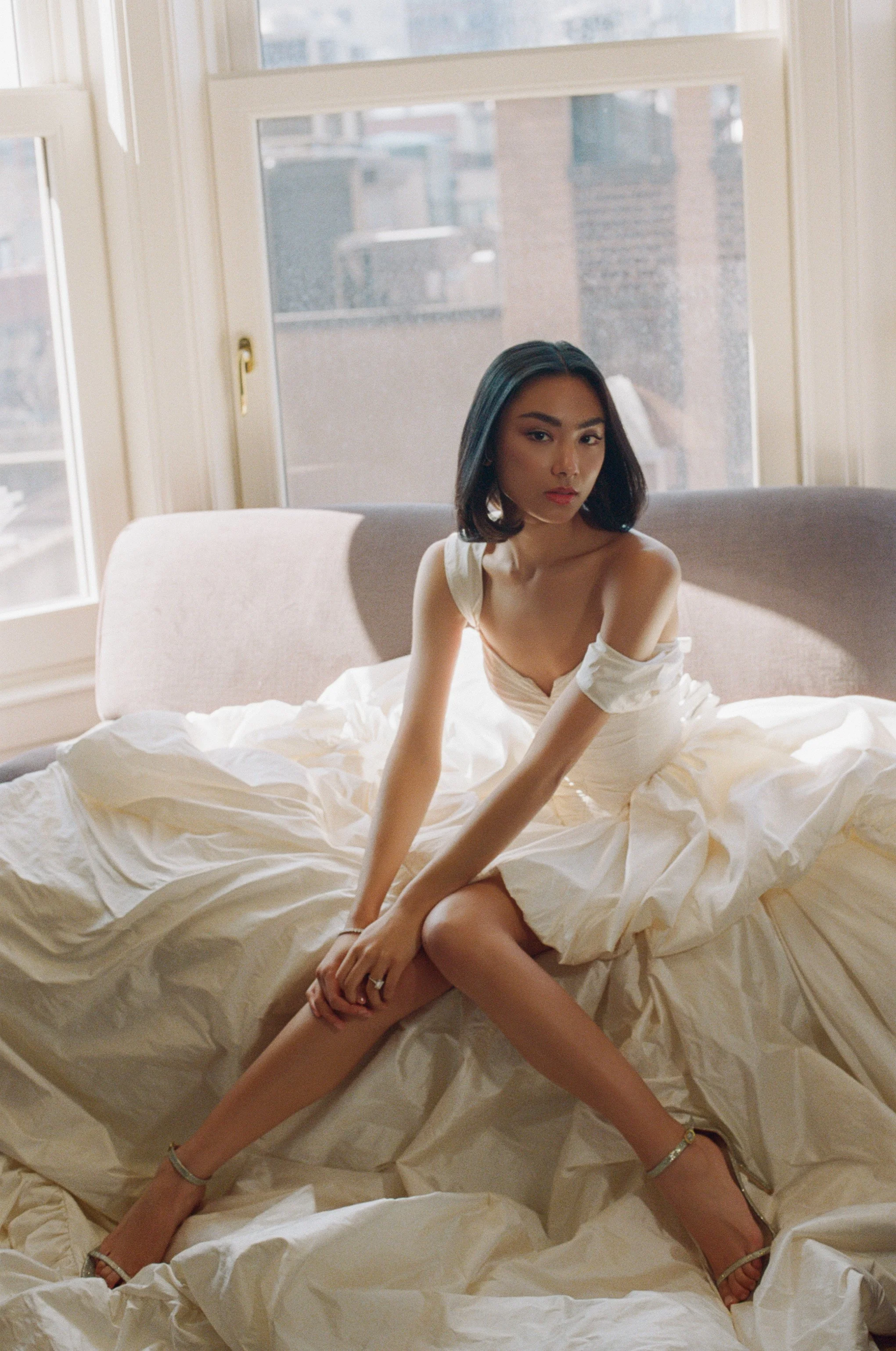 Young woman sitting on a bed with white sheets in a sunlit room, wearing a white dress and high heels.