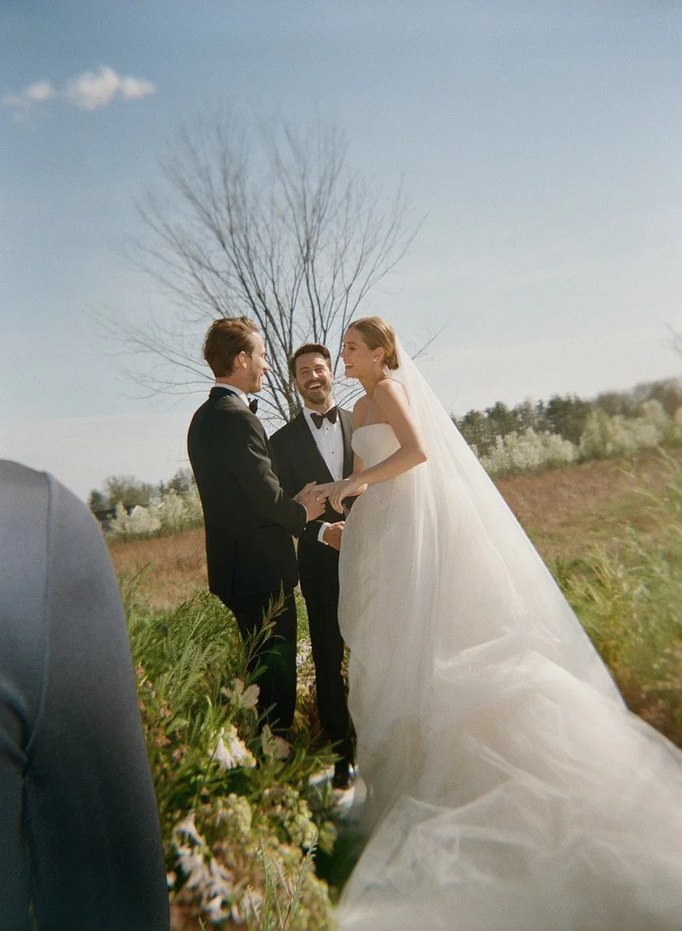 A wedding ceremony outdoors with a bride in a white gown and veil, a groom in a black tuxedo, and an officiant, with a large tree in the background and a blue sky.