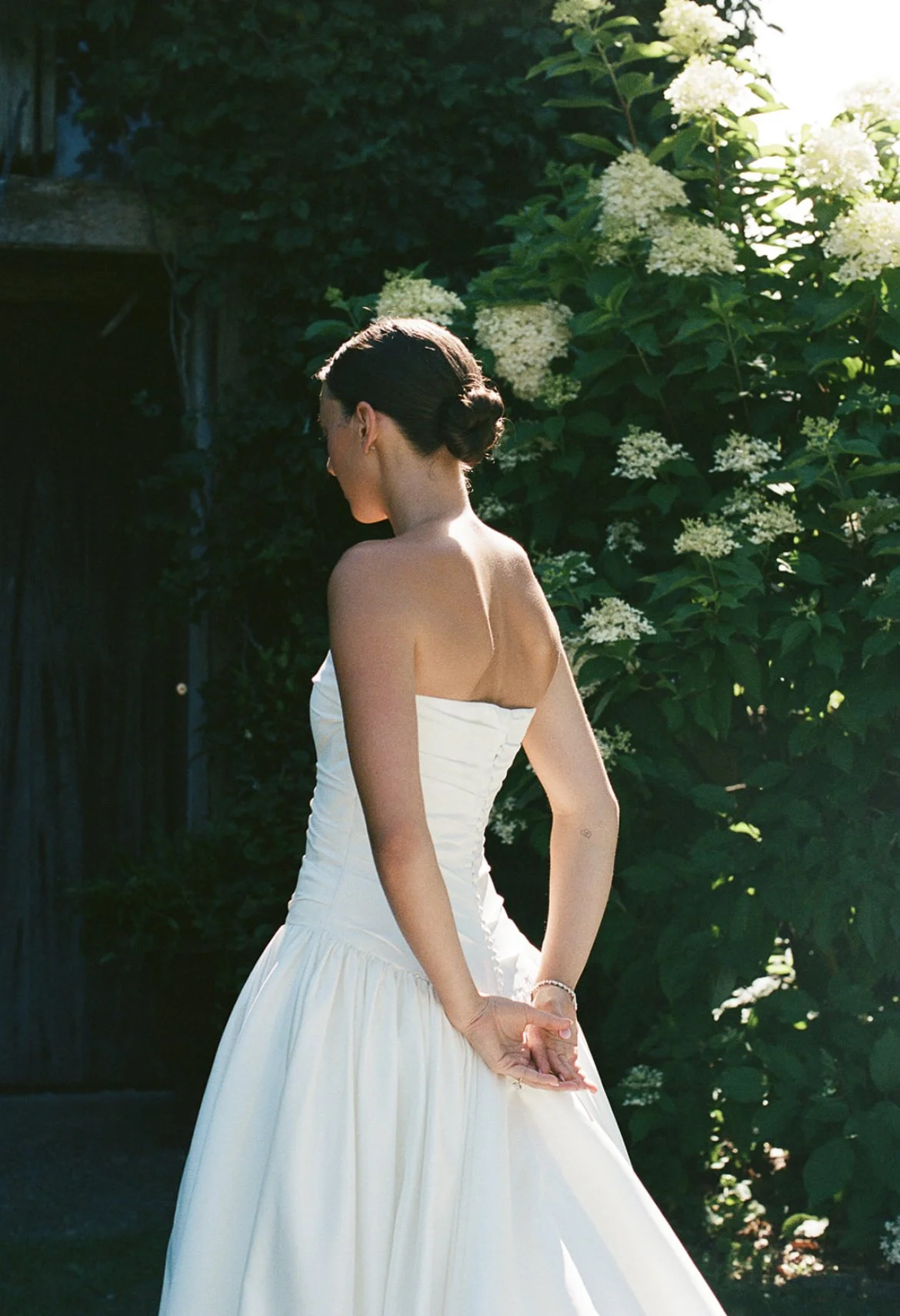 A woman in a strapless white Danielle Frankel wedding dress stands outdoors with her back partially turned, hands clasped behind her back. She is surrounded by green foliage and white flowers, with sunlight illuminating her.