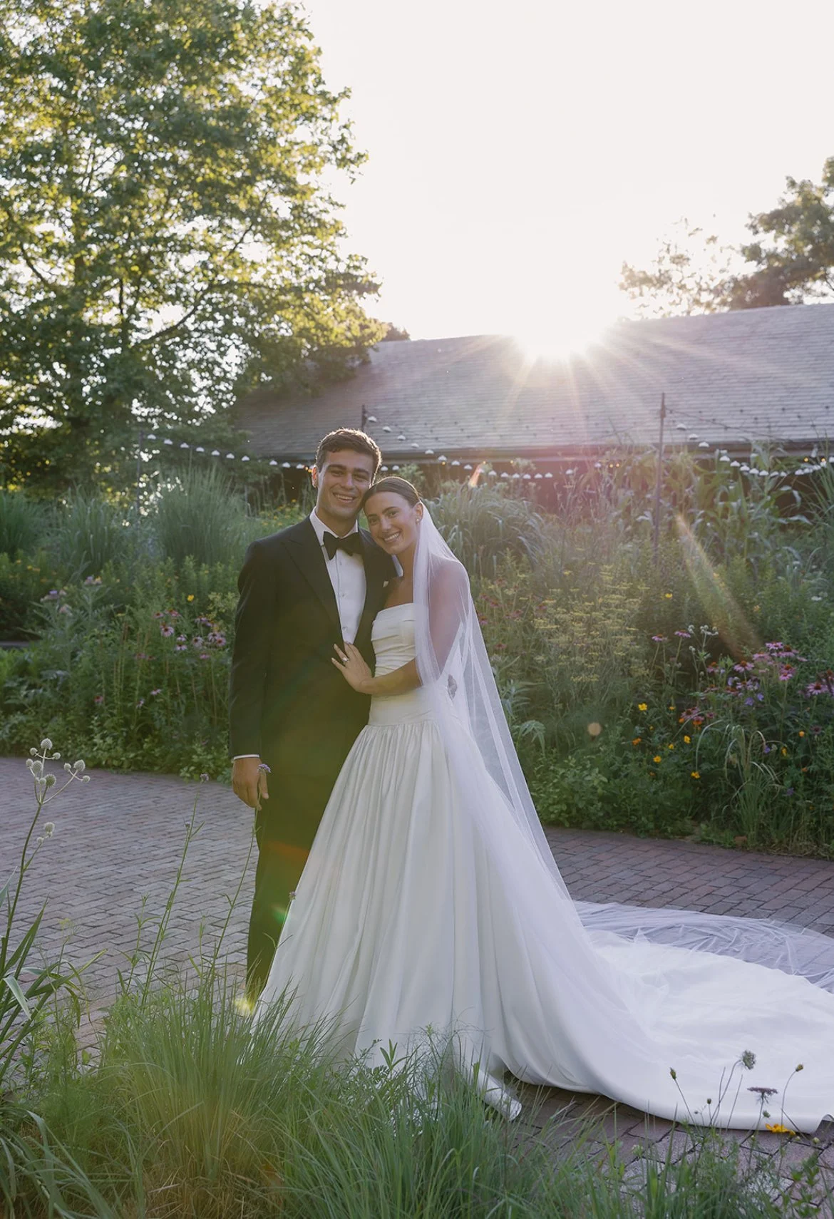 A newlywed couple stands together outdoors during sunset, smiling. The groom wears a black tuxedo with a bow tie, and the bride wears a white wedding gown with a long veil. They are surrounded by greenery and flowers, with the sun shining brightly in