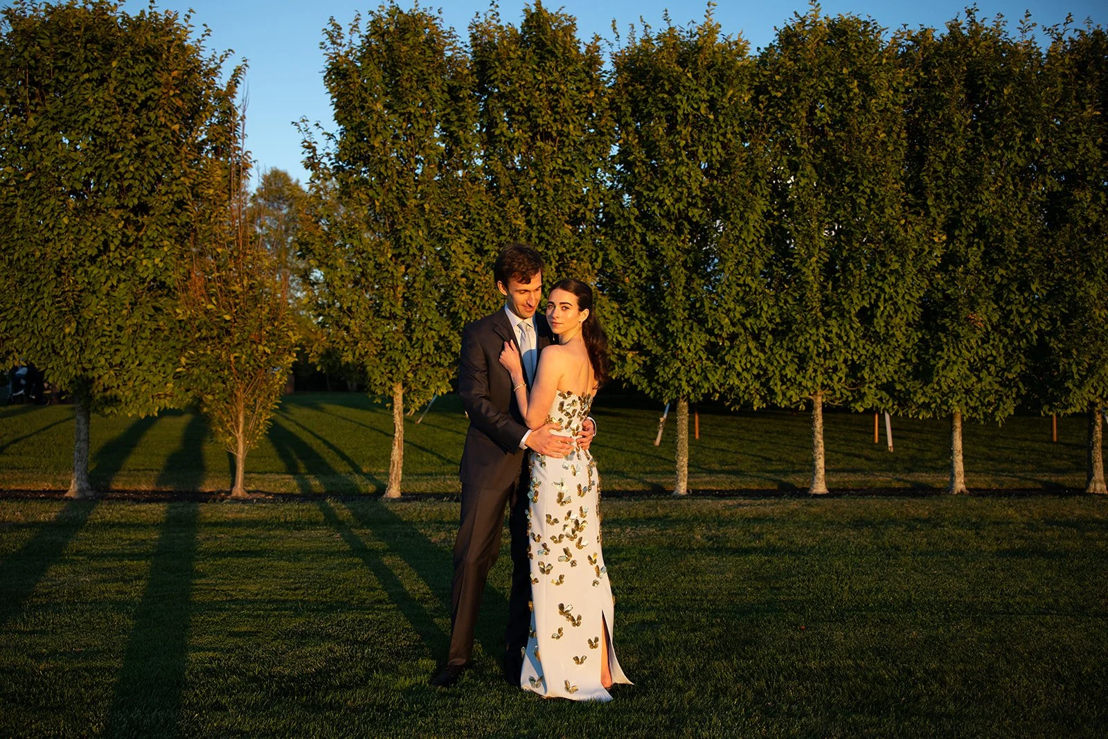 A couple dressed in formal attire stands closely together on a grassy field with tall trees in the background during sunset.