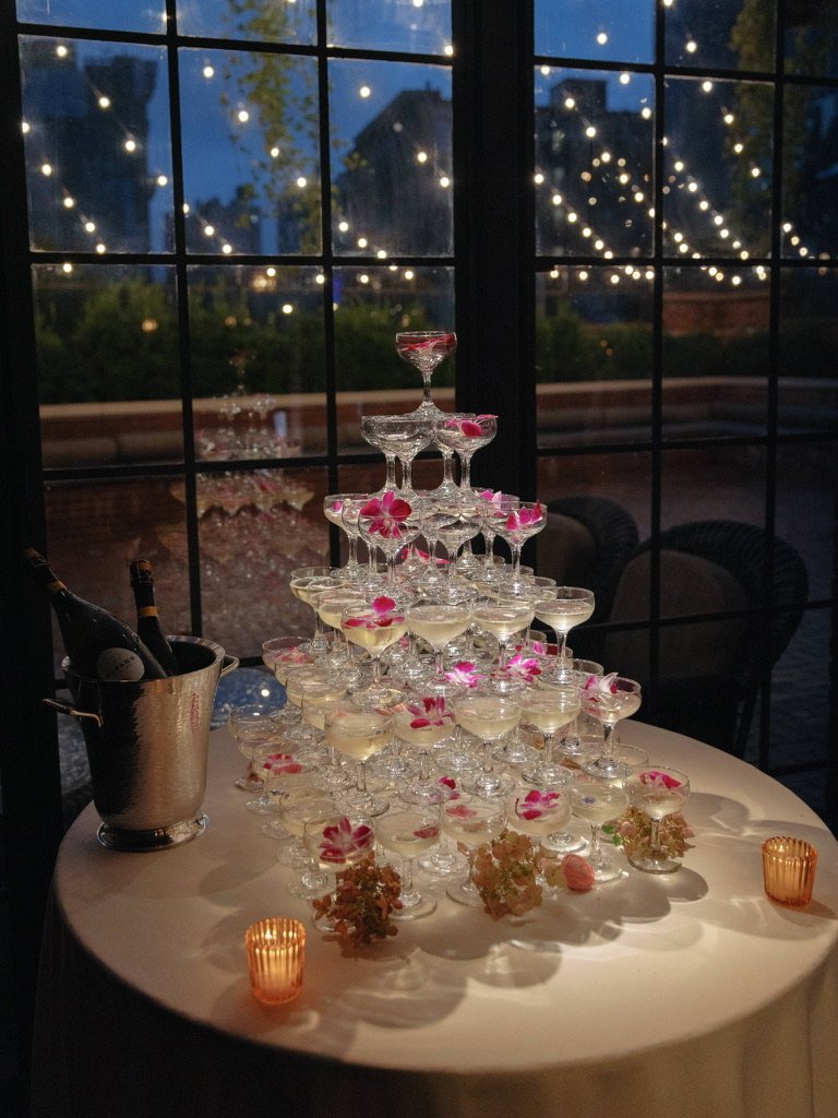 A pyramid of champagne glasses decorated with pink flowers on a table, with candle holders and a bucket of champagne bottles, against a window with string lights outside.