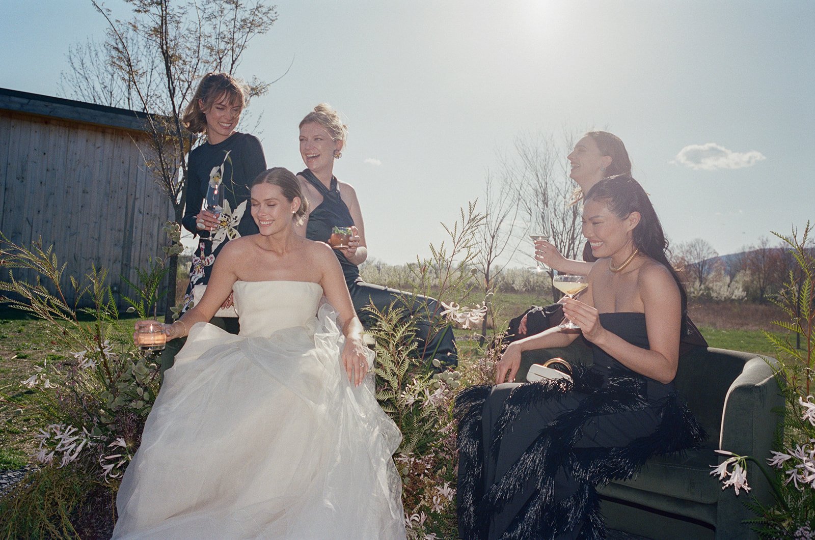 Six women dressed in elegant black dresses and one in a white wedding gown, enjoying drinks outdoors on a sunny day with a wooden building and trees in the background.
