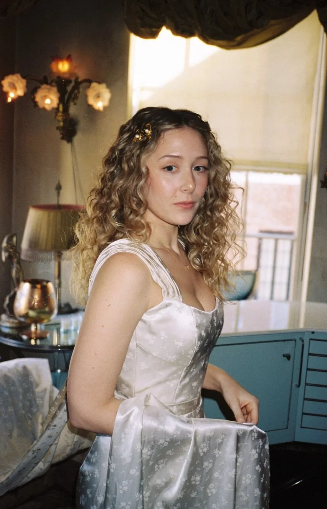 A woman with curly blonde hair wearing a vintage white satin dress. She is standing in a room with retro decor, including a turquoise cabinet, a table lamp, and a wall sconce with pink flowers.