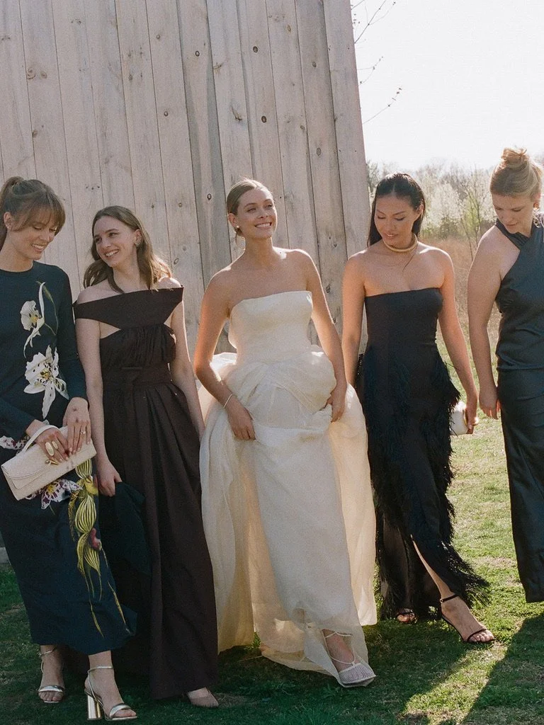 Group of five women dressed in formal attire, standing outdoors in front of a wooden wall, smiling and walking together.