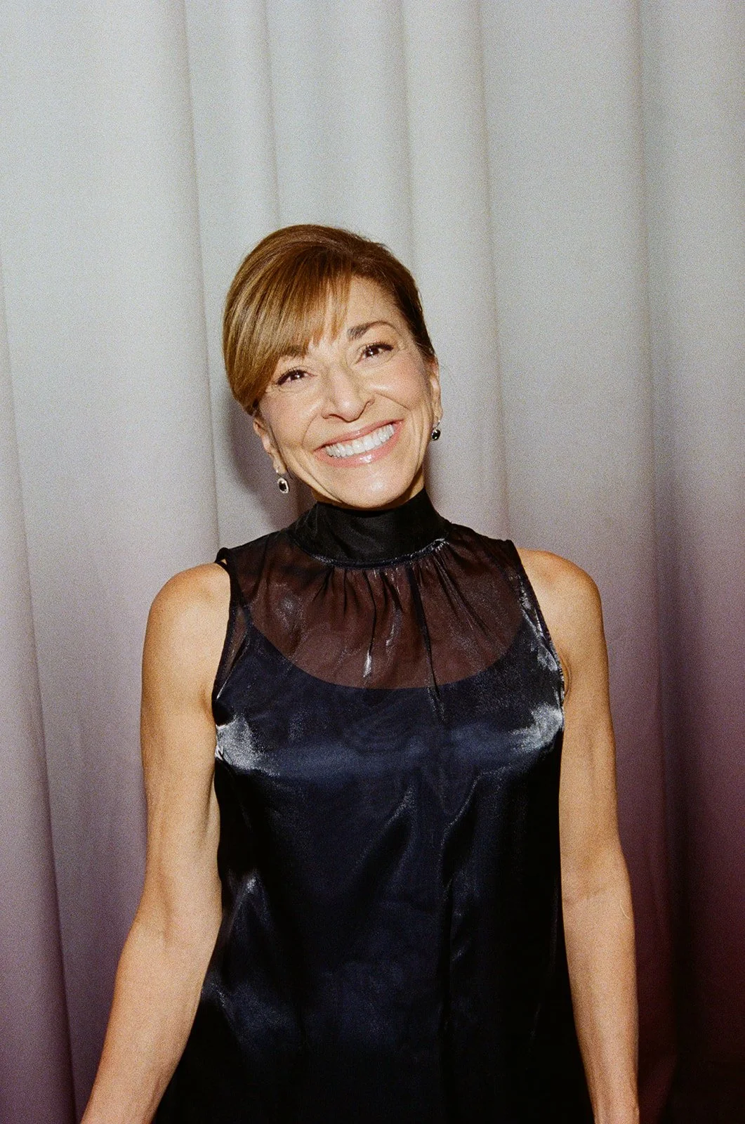 A woman with short brown hair in a sleeveless black dress, smiling in front of a plain background.