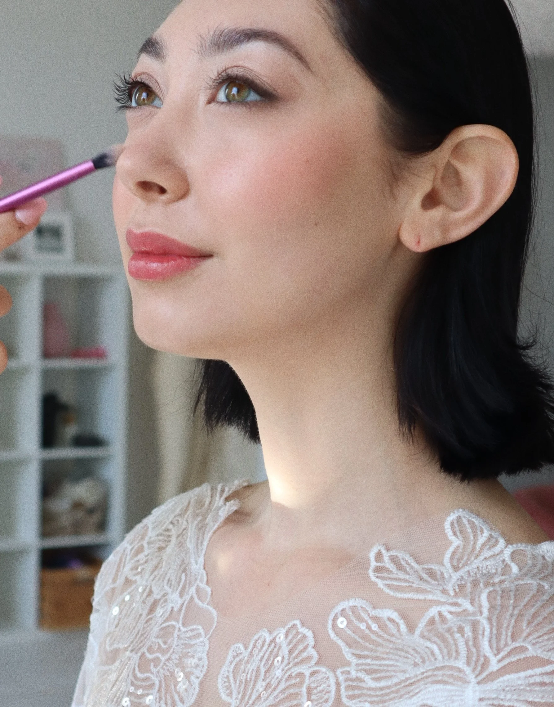 A woman having makeup applied to her face with a brush, wearing a delicate lace top, with a calm expression and standing in a softly lit room.