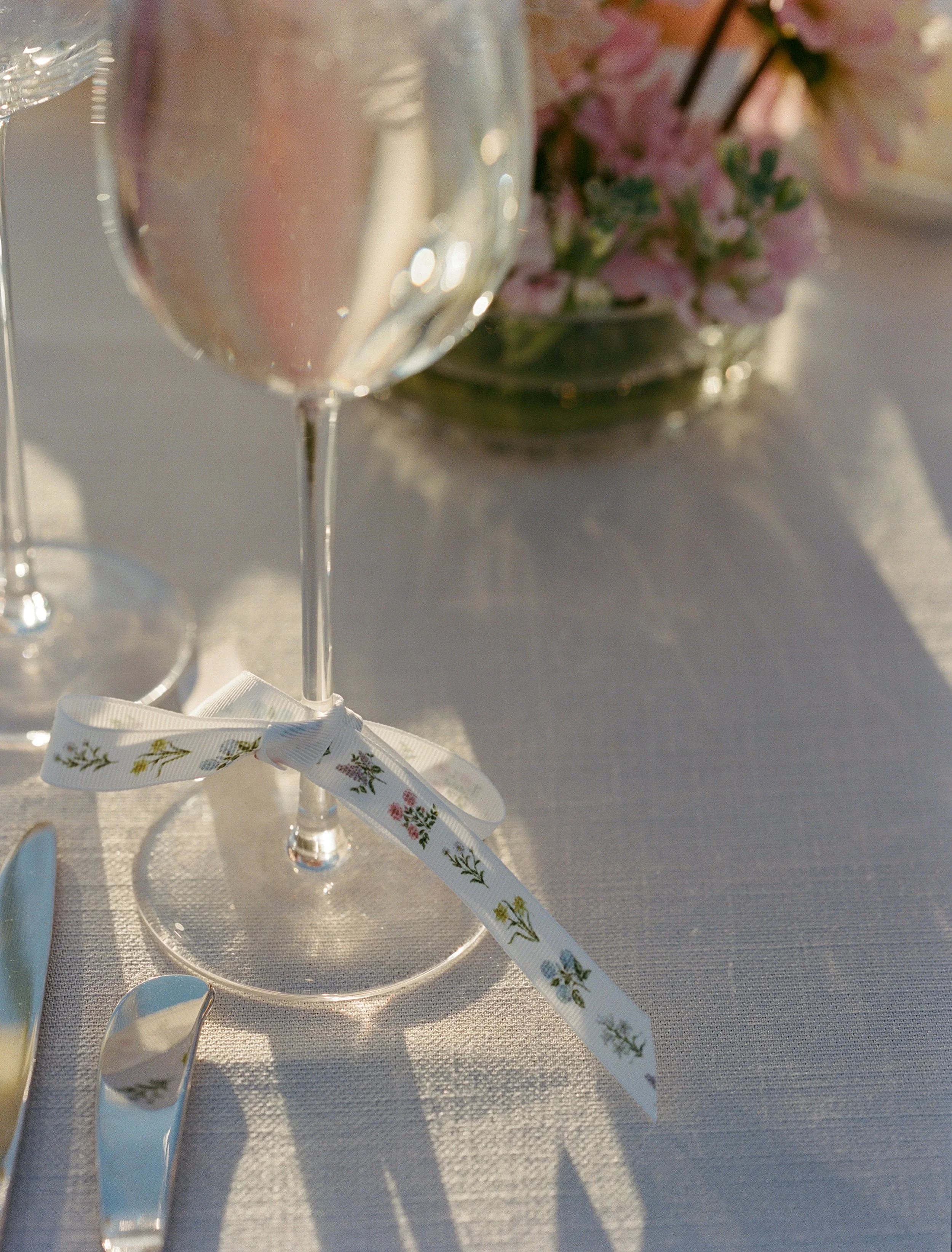 Close-up of a wine glass with a ribbon tied around its stem, set on a table with silverware and a flower arrangement in the background.